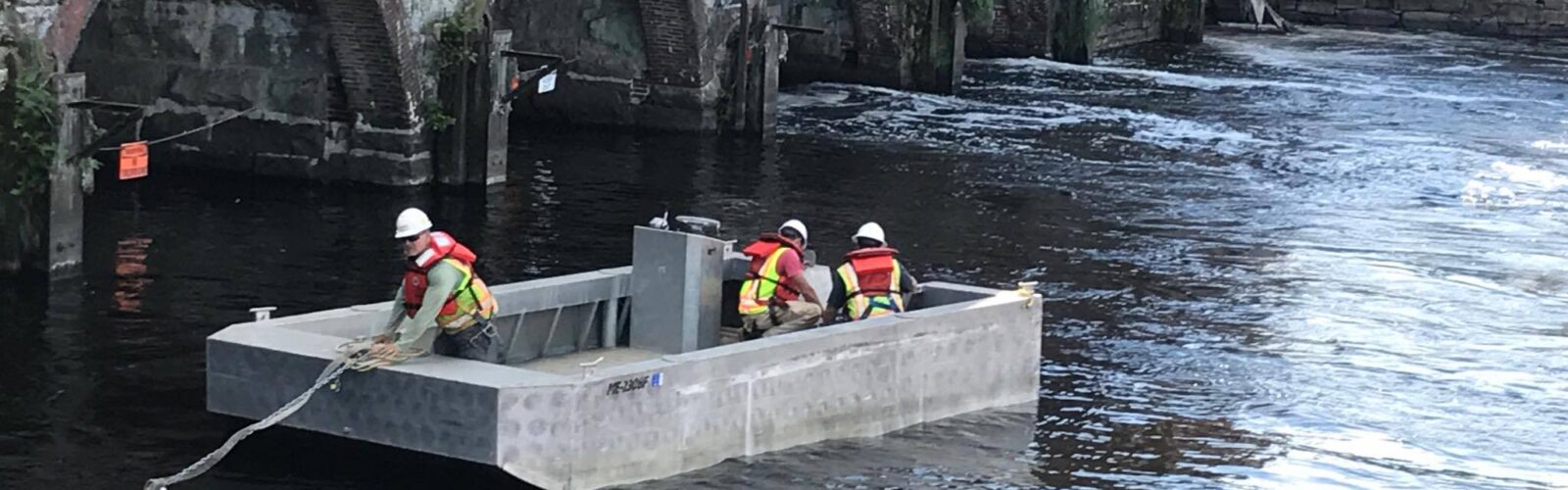 photo of three engineers in a square boat on a river next to a building